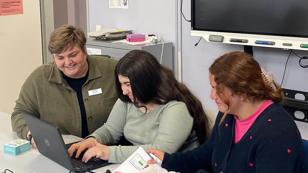 Drei Frauen sitzen gemeinsam an einem Tisch in einem Klassenraum. Eine Frau lächelt und schaut auf den Laptop-Bildschirm, während eine zweite Frau tippt. Die dritte Frau hält ein aufgeschlagenes Buch und zeigt auf eine Seite. Im Hintergrund ist ein Smartboard zu sehen.